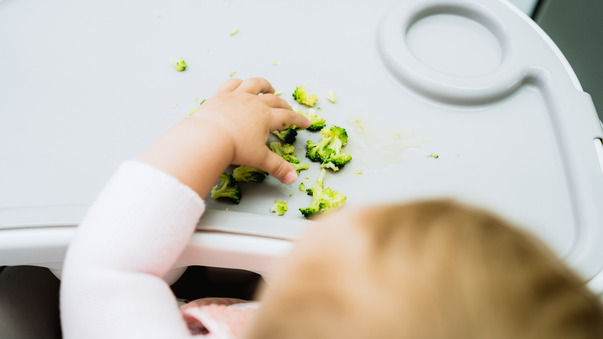 Image looking down on top of infant's head with their hand reaching out to pick up some broccoli off a high chair